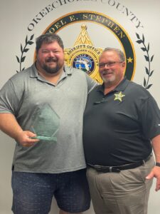 Philip DeBerard on left in a grey polo holding a clear award. Sheriff Noel E. Stephen on the right in a black polo with the Okeechobee County Sheriff's Department logo in the background.